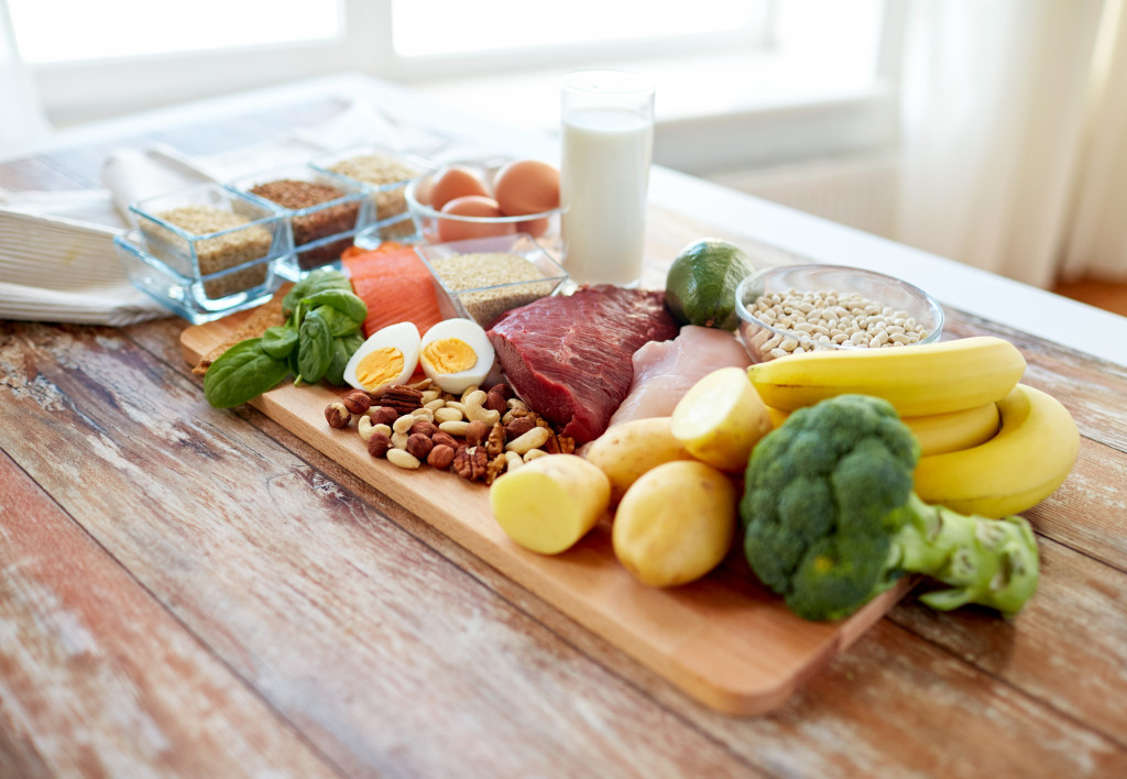 Wooden cutting board with fruits, vegetables, meat, bread, and nuts placed on a table with a glass of milk and eggs on the side.